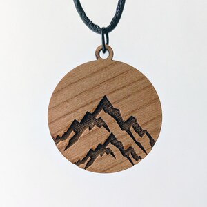 May include: A round wooden pendant necklace with a mountain range design. The pendant is made of light brown wood and features a carved silhouette of three mountain peaks. The necklace has a black cord.