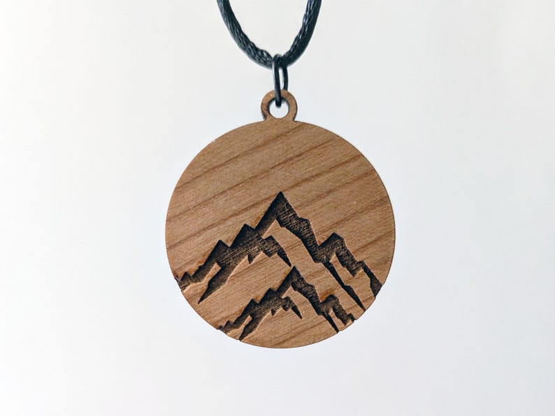 May include: A round wooden pendant necklace with a mountain range design. The pendant is made of light brown wood and features a carved silhouette of three mountain peaks. The necklace has a black cord.