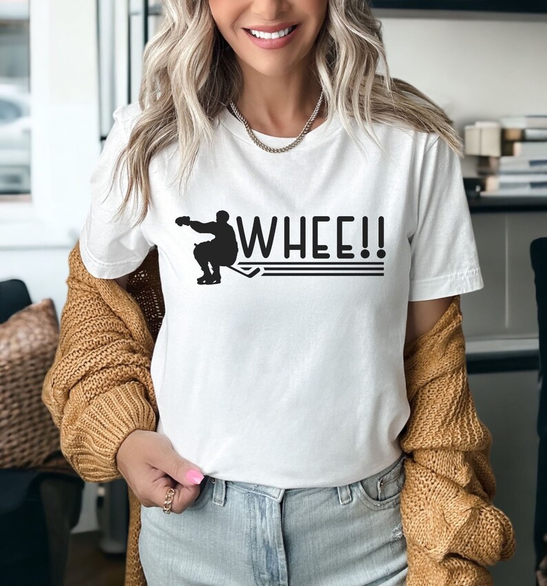 K&ouml;nnte beinhalten: Wei&szlig;es T-Shirt mit einer schwarzen Silhouette eines Hockeyspielers und dem Text "WHEE!!" auf der Vorderseite.