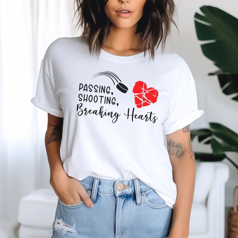 K&ouml;nnte beinhalten: Wei&szlig;es T-Shirt mit einem schwarzen und roten Motiv. Das Motiv zeigt einen Hockeypuck, der durch die Luft fliegt, und ein gebrochenes Herz. Der Text auf dem T-Shirt lautet "Passing, Shooting, Breaking Hearts."