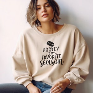 K&ouml;nnte beinhalten: Ein beiger Pullover mit dem Text "Hockey is my favorite season" und einer Grafik eines Hockeyschl&auml;gers.