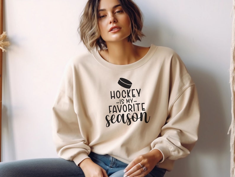 K&ouml;nnte beinhalten: Ein beiger Pullover mit dem Text "Hockey is my favorite season" und einer Grafik eines Hockeyschl&auml;gers.