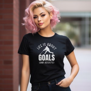 K&ouml;nnte beinhalten: Schwarzes T-Shirt mit einer wei&szlig;en Grafik eines Hockeyspielers und dem Text "Life is about goals (and assists)".