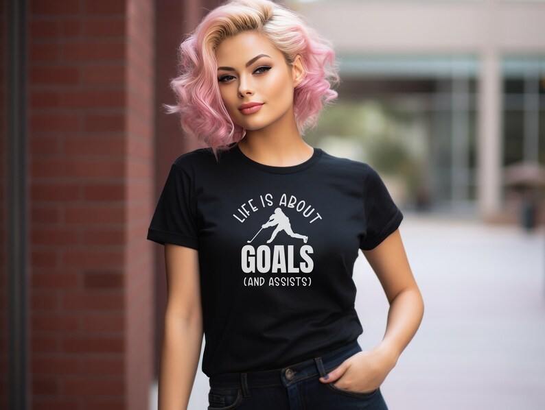 K&ouml;nnte beinhalten: Schwarzes T-Shirt mit einer wei&szlig;en Grafik eines Hockeyspielers und dem Text "Life is about goals (and assists)".