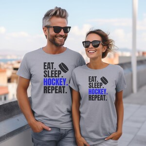 K&ouml;nnte beinhalten: Zwei graue T-Shirts mit dem Text "Eat. Sleep. Hockey. Repeat." und einer blauen Hockey-Puck-Grafik. Die Shirts werden von zwei Personen getragen, die im Freien stehen.