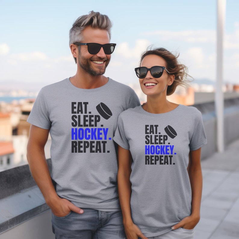 K&ouml;nnte beinhalten: Zwei graue T-Shirts mit dem Text "Eat. Sleep. Hockey. Repeat." und einer blauen Hockey-Puck-Grafik. Die Shirts werden von zwei Personen getragen, die im Freien stehen.