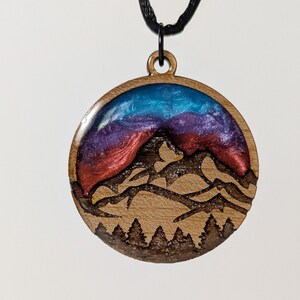May include: Wooden pendant with a mountain scene. The round pendant features a mountain range design with a blue, purple, and red resin sky. The pendant hangs from a black cord.