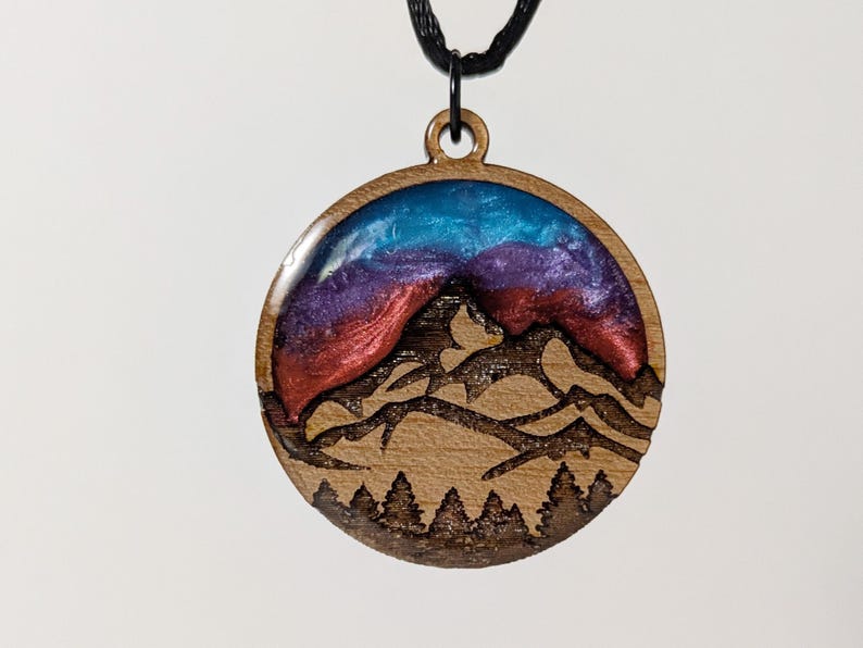 May include: Wooden pendant with a mountain scene. The round pendant features a mountain range design with a blue, purple, and red resin sky. The pendant hangs from a black cord.