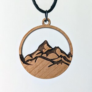 May include: A wooden pendant necklace featuring a mountain range design. The pendant is a circular shape with a mountain silhouette carved inside. The necklace has a black cord.