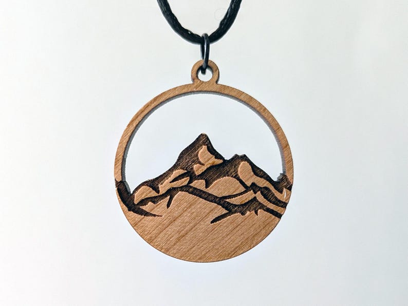 May include: A wooden pendant necklace featuring a mountain range design. The pendant is a circular shape with a mountain silhouette carved inside. The necklace has a black cord.
