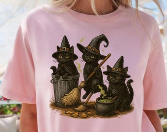 May include: Light pink t-shirt featuring a graphic of three black cats wearing witch hats. One cat is in a trash can, another is holding a broom, and the third is near a cauldron. The design has a Halloween theme.