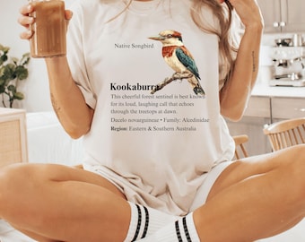 Puede incluir: Camiseta blanca con una ilustración colorida de un pájaro Kookaburra, con el texto "Native Songbird" e información sobre el ave. La camiseta se combina con calcetines blancos con rayas negras. También se ve un vaso con una bebida marrón.