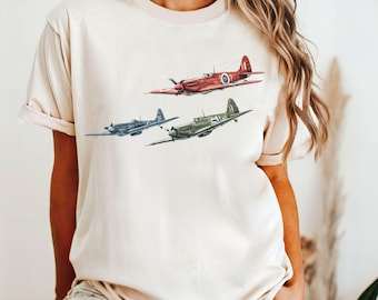 以下が含まれることがあります： クリーム色のTシャツに、3機のビンテージ飛行機のグラフィックがプリントされています。飛行機は赤、緑、青です。画像の右上に「アイボリー」というテキストがあります。