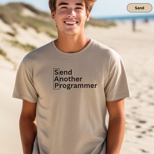 Puede incluir: Una camiseta de manga corta color arena con texto negro en el pecho que dice "Send Another Programmer", con las letras S, A y P alineadas verticalmente en un recuadro negro a la izquierda del resto del texto.