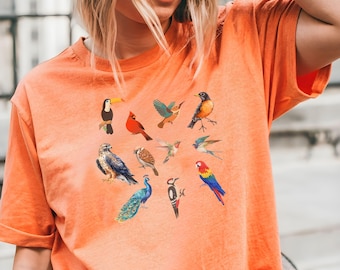 Puede incluir: Una camiseta naranja terracota con un diseño de pájaros coloridos. Los pájaros están dispuestos en forma de corazón e incluyen un tucán, un cardenal, un pavo real, un colibrí y más.