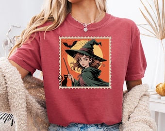 May include: Crimson-colored t-shirt featuring a Halloween-themed graphic. The graphic is a postage stamp design with a witch, black cat, bat, and spiderweb. The witch is wearing a green cape and hat, holding a wand. The shirt is a comfortable, casual style.