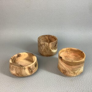May include: Three small, hand-turned wooden bowls with a natural wood grain pattern. The bowls are cylindrical with a rounded interior, showcasing the unique textures and colours of the wood. The bowls are likely for decorative or functional use.