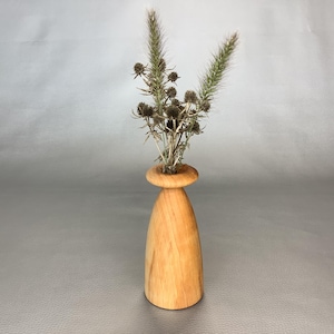 May include: A light-toned wooden bud vase with a smooth, tapered cylindrical body and a flared, rounded rim. The vase contains a small arrangement of dried, spiky brown thistle heads and a single slender, feathery green grass stalk.
