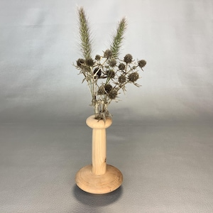 May include: A light-coloured wooden vase with a cylindrical body and a wide, circular base. The vase holds a dried floral arrangement consisting of two tall, feathery green stalks and several small, dark brown, spiky thistle-like flower heads.