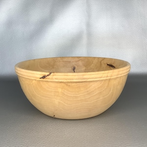 May include: A light-coloured wooden bowl with a smooth, polished finish and visible natural grain patterns. The bowl features a rounded shape with a subtle horizontal ridge near the top rim and small, dark, organic knots embedded in the wood.