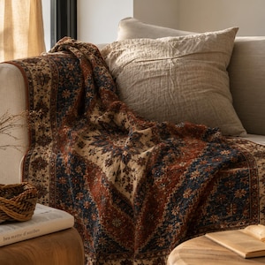 May include: A woven throw blanket featuring a dark blue and rust orange floral pattern with a cream-coloured border. The blanket is draped over a light beige sofa cushion, displaying a detailed, intricate design with repeating motifs and textures.