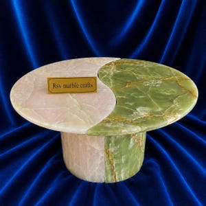 Könnte beinhalten: Ein Couchtisch aus Marmor mit Yin-Yang-Design. Der Tisch besteht aus zwei Farben: Hellrosa und Grün. Der Sockel ist zylindrisch. Der Text "Rsv marble crafts" befindet sich auf einem kleinen goldenen Schild auf dem Tisch.