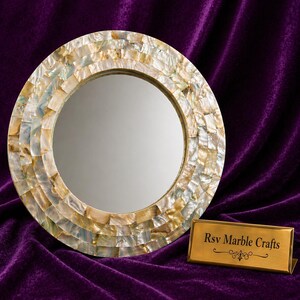 May include: Round decorative mirror with a mosaic shell frame. The frame is made of small, rectangular pieces in shades of cream, gold, and pale blue. A gold-colored plaque with the text "Rsv Marble Crafts" sits below the mirror.