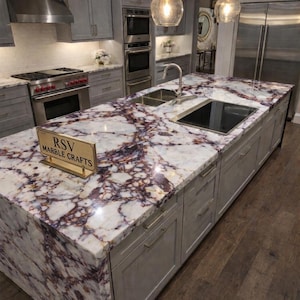 Op de afbeelding: Keuken eiland met een marmeren aanrechtblad met een witte basis met paarse en bruine aderen. Het eiland heeft grijze kasten met zilveren handgrepen. Een bord leest "RSV Marble Crafts".