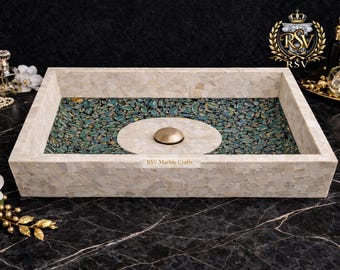 May include: A rectangular decorative tray with a mosaic design. The tray's outer frame is a light beige color, while the interior features a mosaic of teal and green stones. A central, circular element with a gold-colored fixture is present. The text "RSV Marble Crafts" is visible.