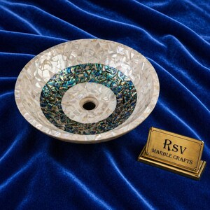 May include: A round, decorative sink with a mosaic design. The sink features a white mother-of-pearl exterior and a central design of blue and green iridescent tiles. A small brass sign with the text "RSV MARBLE CRAFTS" is placed to the side.