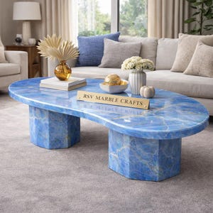 Op de afbeelding: Een blauwe marmeren salontafel met een ovale bovenkant en een geometrische basis. De tafel heeft een opvallende blauwe kleur met witte adering. De tekst "RSV MARBLE CRAFTS" staat op een gouden plaatje.