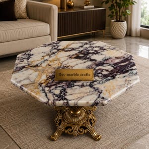 Könnte beinhalten: Ein achteckiger Marmor-Couchtisch mit einem goldfarbenen, verzierten Sockel. Die Tischplatte weist ein weißes, violettes und goldfarbenes Adernmuster auf. Ein goldenes Schild auf dem Tisch trägt die Aufschrift "Rsv marble crafts".