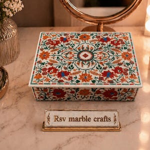 Op de afbeelding: Een rechthoekige witte marmeren doos met een bloemeninlegontwerp in rood, oranje en groen. De doos staat op een marmeren oppervlak, met een klein bordje met de tekst "Rsv marble crafts". Andere items zijn een vaas met bloemen en make-up kwasten.