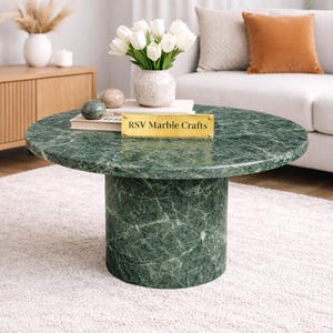 May include: A round, green marble coffee table with a cylindrical base. The table features a polished surface with natural veining. A gold sign on the table reads "RSV Marble Crafts". The table is in a living room setting with a vase of white flowers.