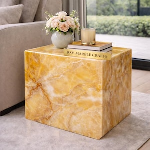 Può includere: Tavolino quadrato effetto marmo giallo dorato con venature marroni. Il tavolo ha una finitura lucida e il testo "RSV MARBLE CRAFTS" è su una placca dorata. Un vaso di fiori, libri e una candela sono sopra.