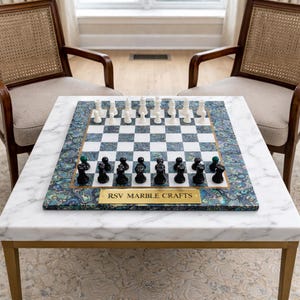 May include: A marble chess set with black and white pieces on a chequered board. The board sits on a white marble table with gold legs. The board has a decorative blue and green border. The text "RSV MARBLE CRAFTS" is on a gold plaque.