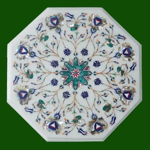 May include: White marble octagonal table with intricate floral inlay design. The table features a central starburst pattern in green and red, surrounded by detailed floral and leaf motifs in various colors. The edges are adorned with silver-colored accents.