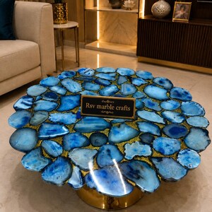 May include: A circular coffee table featuring a tabletop composed of numerous polished blue agate slices arranged in a mosaic pattern with gold-colored resin filling the gaps. A black rectangular sign with the text Rsv marble crafts sits on top.