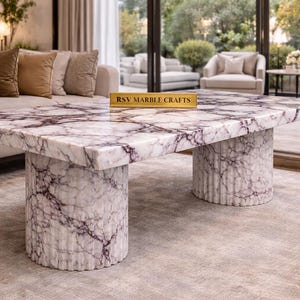 Könnte beinhalten: Ein quadratischer Marmor-Couchtisch mit einem weiß-violett geäderten Muster. Der Tisch hat zwei zylindrische Sockel. Der Text "RSV MARBLE CRAFTS" befindet sich auf einem kleinen goldenen Schild auf dem Tisch. Der Tisch befindet sich in einem Wohnzimmer.