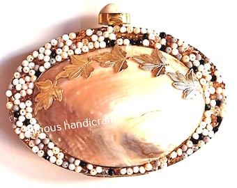 May include: A beige oval clutch purse with a shell design and gold leaf accents. The purse is decorated with white, black, and gold beads.