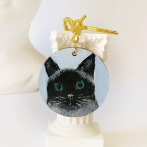 Può includere: Un portachiavi rotondo in legno con un gatto nero con occhi blu brillanti. Il portachiavi è attaccato a un anello di metallo dorato.