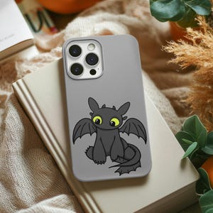 Può includere: Una custodia per telefono grigia con un design a cartone animato di un drago, con grandi occhi gialli e ali nere. La custodia è appoggiata su un libro chiuso. Lo sfondo include un tessuto beige morbido ed elementi decorativi.