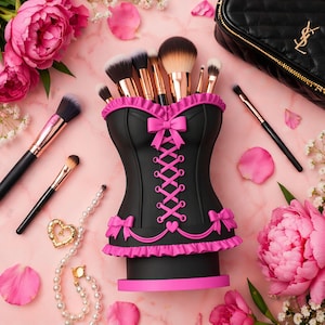 Può includere: Un porta pennelli per trucco a forma di corsetto nero e rosa con un design allacciato, contenente vari pennelli per trucco. Il supporto è circondato da fiori rosa, pennelli per trucco, una collana di perle e una borsa nera.