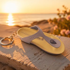 Può includere: Un portachiavi a infradito in miniatura con suola gialla, cinturini grigi e un portachiavi argentato. Il portachiavi è appoggiato su una superficie strutturata con uno sfondo sfocato dell'oceano e del tramonto.
