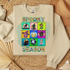 Può includere: Felpa beige con una grafica colorata a tema Halloween. La grafica presenta le parole "Spooky Season" e nove immagini quadrate di icone di Halloween, tra cui un gatto nero, un gufo e una zucca. Il design è delineato da un bordo decorativo.