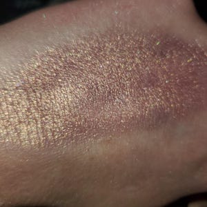 May include: Close-up of a skin surface with a shimmering, iridescent makeup application. The product appears to be a blend of gold, pink, and bronze tones, with a glittery, textured finish. The light catches the product, creating a sparkling effect.