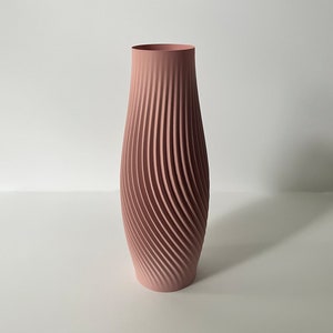 Pode incluir: Um vaso alto, cor de rosa, com um design canelado em espiral. O vaso tem uma base larga que se estreita para um pescoço estreito e uma borda lisa. As nervuras verticais criam uma superfície texturizada. O vaso é feito de material sólido.