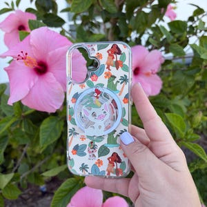 May include: Clear phone case with a colorful design featuring cartoon characters, palm trees, and flowers. The case has a pop-up grip with a cartoon pig design. The background includes pink hibiscus flowers and green foliage.