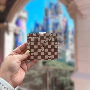 Op de afbeelding: Een bruin-wit geblokte kaarthouder met een geschulpte bovenrand en een drukknoopsluiting. De kaarthouder heeft Mickey Mouse silhouetten en een goudkleurige sleutelhanger. De achtergrond is wazig, met een kasteel zichtbaar.