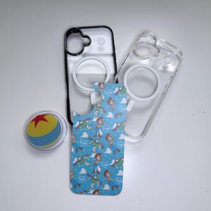 May include: A collection of phone accessories, including two clear phone cases, one with a black border, and a blue patterned insert featuring cartoon characters. A round, clear phone grip with a yellow, red, and blue ball design is also present.